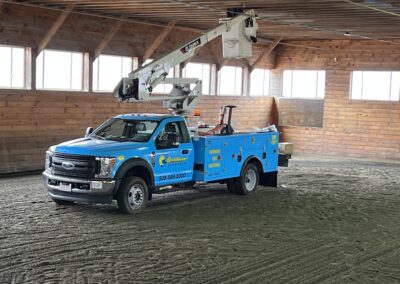 A blue utility truck with an extended boom lift parked inside a wooden structure. The truck has company signage and contact information on the side.