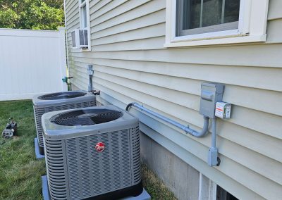 Two air conditioning units are installed outside a beige house, with wiring and conduits running along the siding near windows.