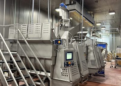 Industrial food processing machine with control panel and metal stairs in a large, well-lit facility with concrete floors and overhead lighting.