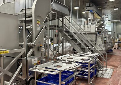 Industrial food processing equipment with metal conveyors and blue crates inside a large, clean facility with concrete floors and high ceilings.
