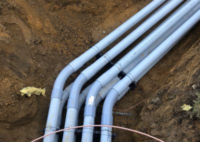 Five gray PVC pipes run underground through a dirt trench, secured with black fasteners, next to a section of exposed rebar and a piece of yellow debris.