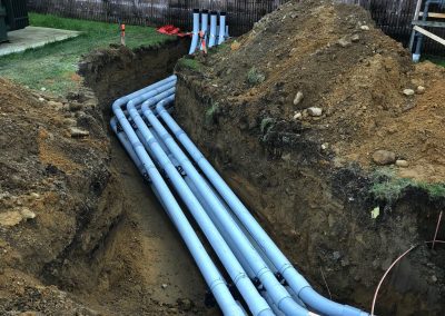 A trench with multiple gray PVC conduits installed underground next to a building with a red wall. Dirt piles are on both sides of the trench.