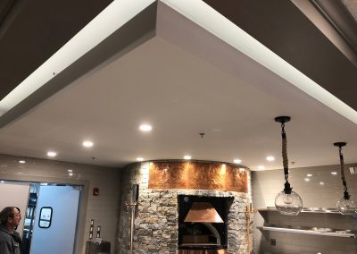 Modern commercial kitchen with stone oven, copper hood, pendant lights, white tile walls, and a person standing near a door.