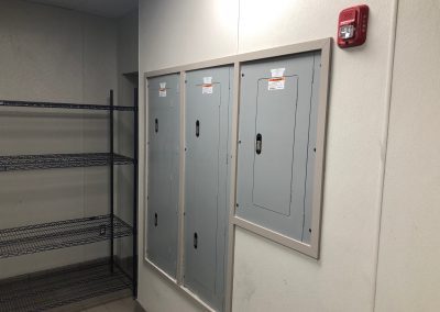 Three gray electrical panels on a wall next to a metal shelving unit; a red fire alarm is mounted above the panels.