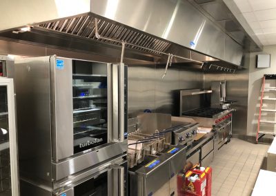 Commercial kitchen with ovens, stoves, deep fryers, stainless steel surfaces, and storage racks. A red fire extinguisher and a step ladder are visible on the right side.