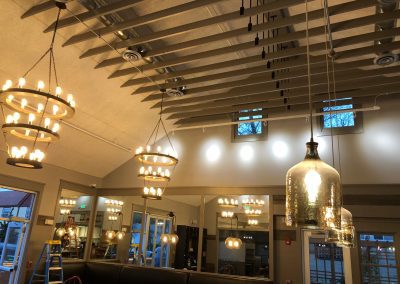 Modern restaurant interior with high ceiling, exposed beams, multiple chandeliers, pendant lights, large windows, and mirrors reflecting the lighting.