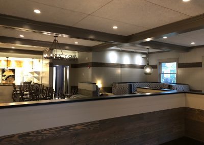 Empty restaurant interior with stacked chairs on tables, empty booths, pendant lighting, and a sign indicating restrooms in the background.