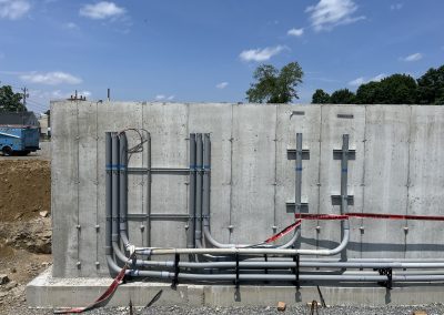 Gray electrical conduits are mounted and run along a concrete wall at a construction site under a sunny sky, with some dirt and a utility truck visible in the background.