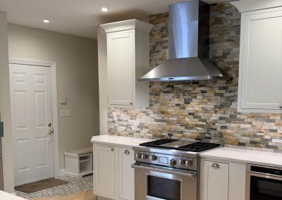 Modern kitchen with stainless steel stove and vent hood, stone tile backsplash, white cabinets, and light wood floor. A door and small tiled area are visible in the background.