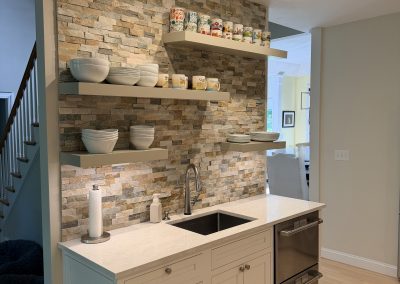 Modern kitchen with stone backsplash, open shelves holding dishes and mugs, stainless sink, and under-counter appliances. Bright overhead lighting highlights the clean, organized space.