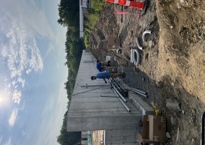 Two workers in blue shirts install electrical conduit next to a concrete wall at a construction site, with boxes, pipes, and orange traffic cones nearby under a partly cloudy sky.
